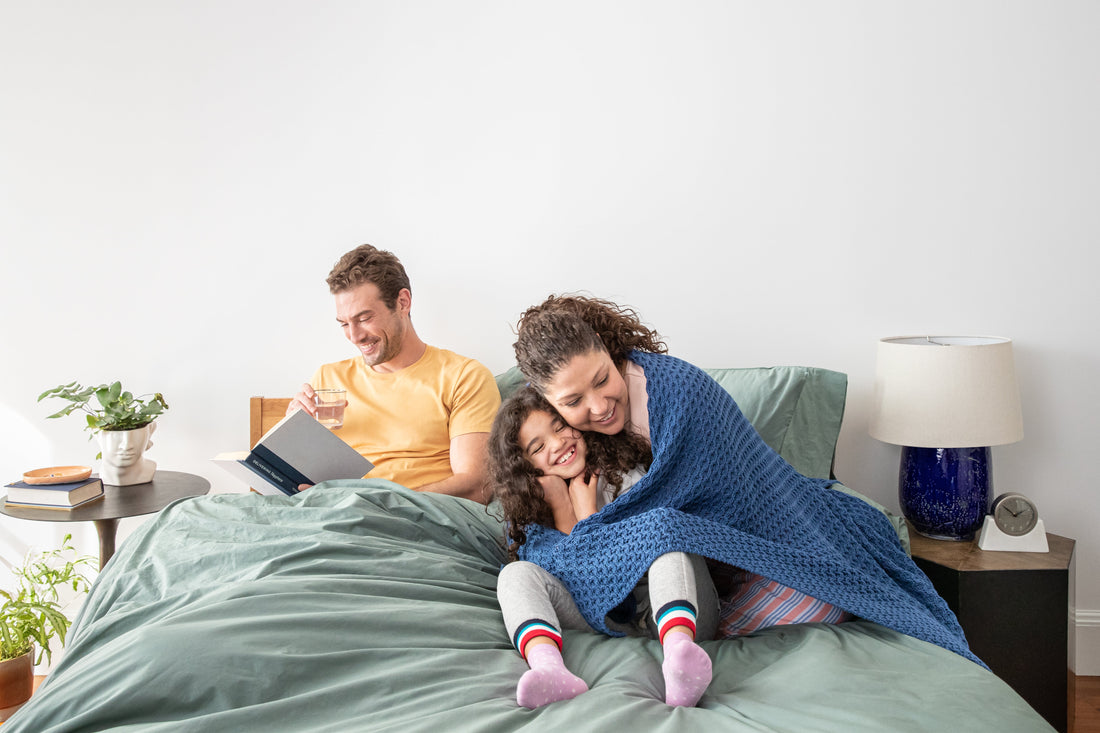 Family of three cuddling, reading and laughing in a bed made with sage colored bedding