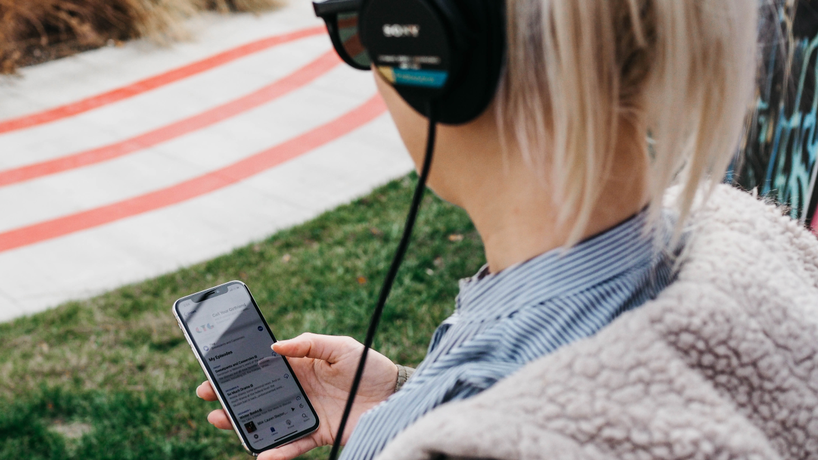 a woman wearing black headphones and holding a smartphone in her hand. The picture was taken from the perspective of someone looking over her shoulder.