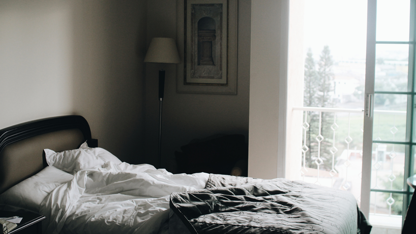 Picture of a bed that looks slept in with white sheets and a grey blanket with an open window in the background and sun coming thru it