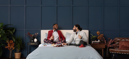 Two men in formal attire laughing and drinking champagne on top of a bed.