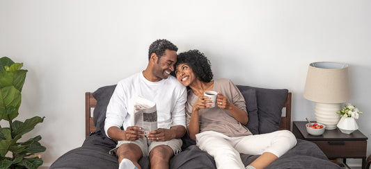 Couple sitting up in bed next to each other smiling while reading and holding a coffee mug