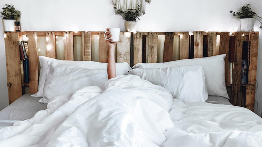 Someone laying in bed under white covers raising only their hand up that is holding a coffee mug.