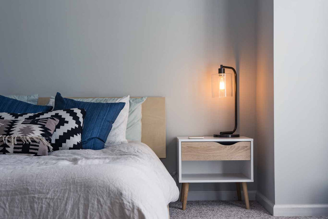Picture showing half of a bed made with grey and blue bedding with a nightstand next to the bed with a light sitting on top of it