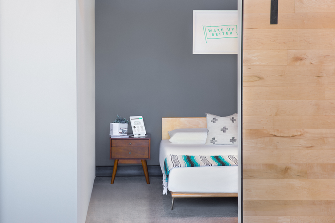 Looking thru a doorway at a bed made with white bedding and colorful blanket at the foot of the bed and nightstand next to it