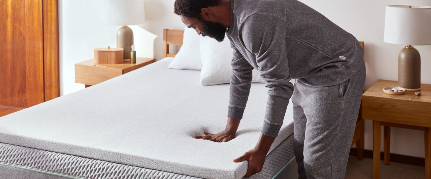 Man pressing his left hand on a great mattress that has two white pillows on.  Pressing shows the softness of the mattress.  The mattress is on a brown bed frame with 2 end tables on both side.