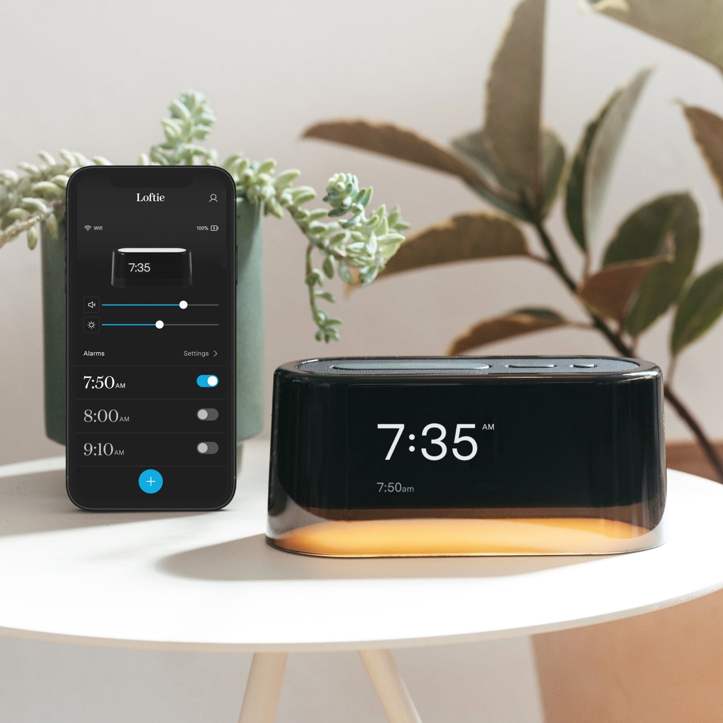 The Tuft and Needle Loftie Smart Alarm Clock in a bedroom on a table next to a phone with the Loftie app