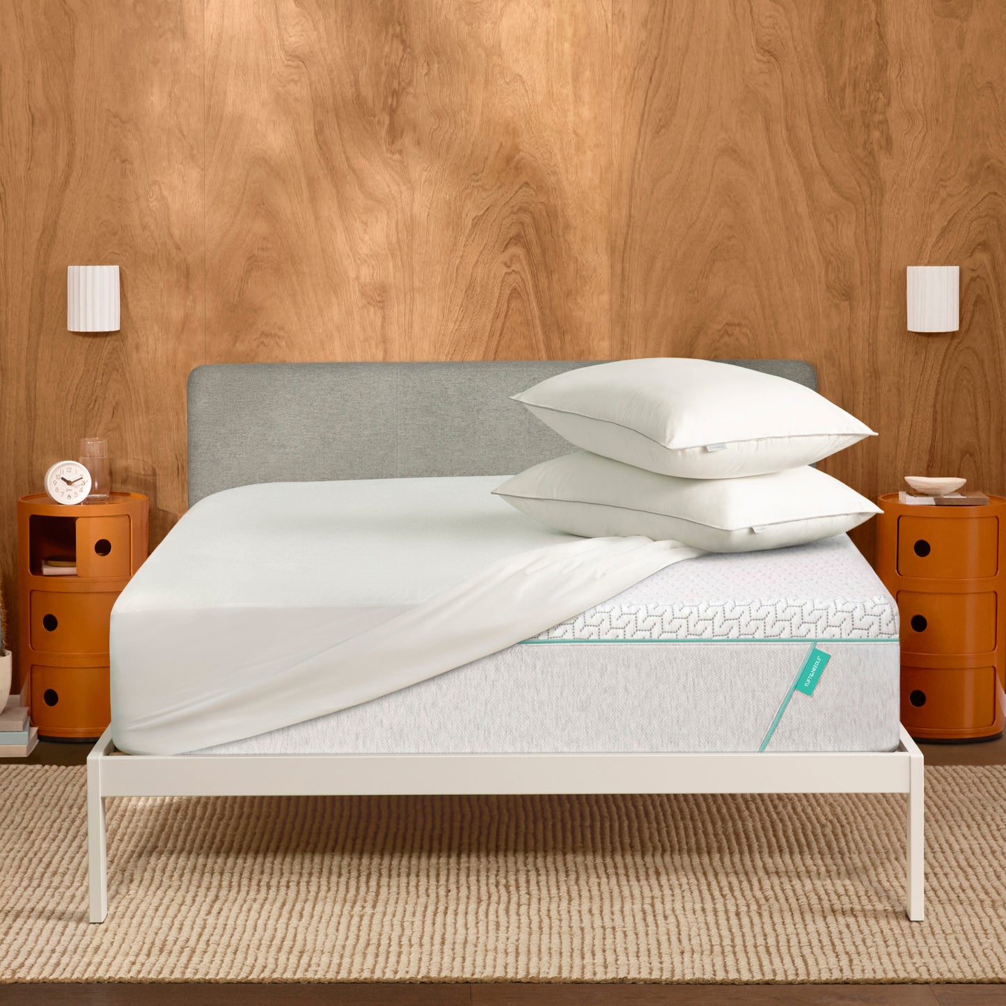 Tuft & Needle Bed with white mattress, gray headboard, and white pillows against a wooden wall.