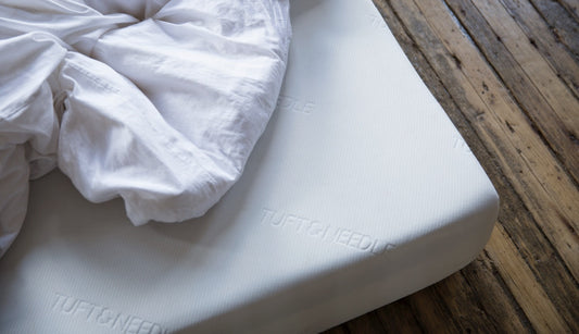 Corner of an Original Tuft & Needle Mattress sitting on a wood floor with white sheets piled on top of it