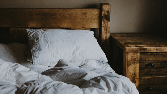 A white pillow and white bedding looking slept in on a bed with a dark wood bed frame and matching night stand next to it