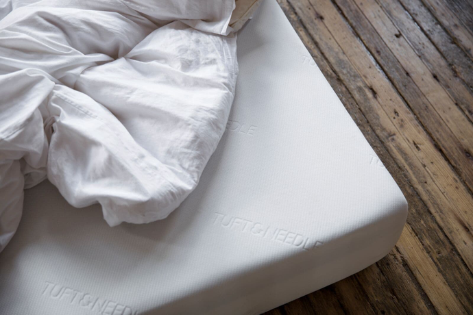 Picture of the corner of a Tuft & Needle Original Mattress sitting on a wood floor with white sheets piled on top of it