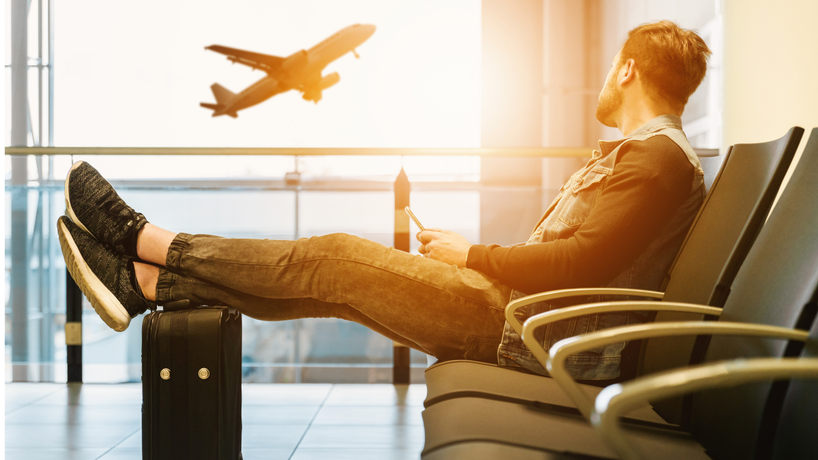 Man sitting in an airport terminal with his feet on his luggage looking out the window at a plane taking off