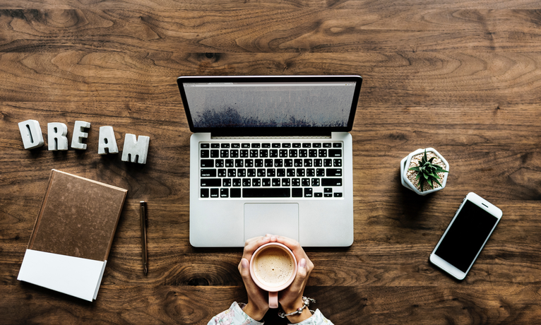 Overhead view of a desk with a laptop, phone, small plant, journal, a cup of coffee and the word dream on it