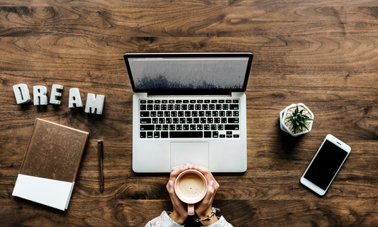 Overhead view of a desk with a laptop, phone, small plant, journal, a cup of coffee and the word dream on it