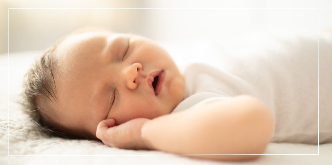 Picture of a baby sleeping with its hand up by its face and its mouth open. Baby is in a white onsie.