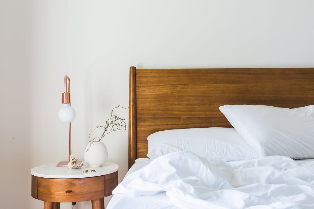 Corner of a bed with a dark wood headboard made with white bedding and a round night stand with a gold lamp and little plant sitting next to it.