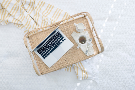 Arial photo of a laptop opened and a cup of tea and cream sitting next to it, all on a light wood tray