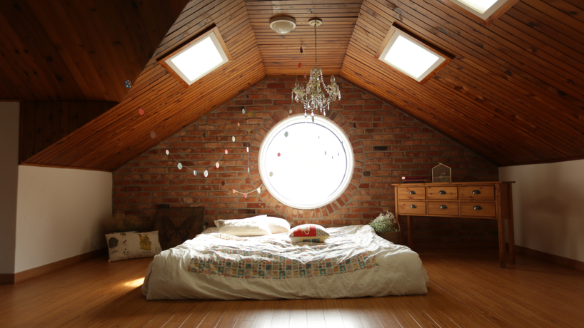 Picture of a mattress on the floor of a triangular shaped loft with a circle window above it and some sky lights