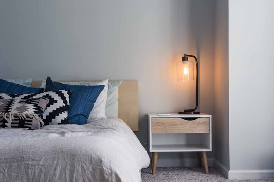 Picture showing half of a bed made with grey and blue bedding with a nightstand next to the bed with a light sitting on top of it