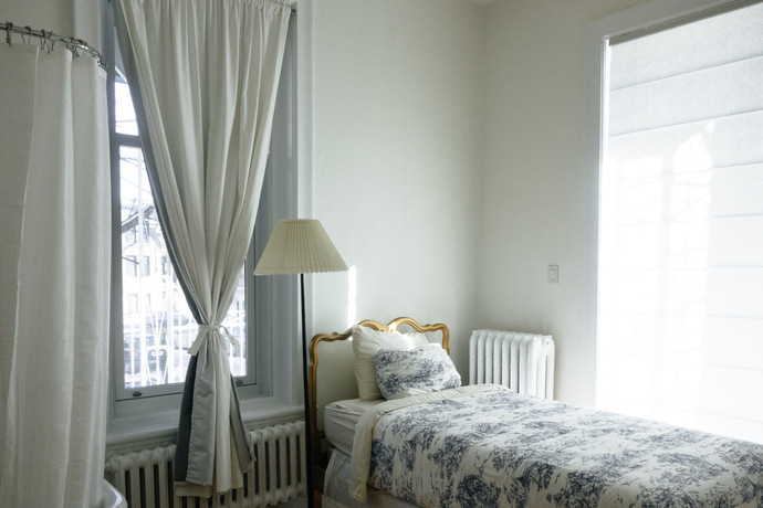 Twin bed in corner of a room with a lamp next to it and two windows. One covered by sheer window blinds and the other covered by a white curtain tied in the middle and draping down.