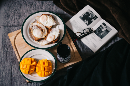 Picture of a breakfast with eggs and mangos on a tray sitting on a bed with a book opened up and glasses sitting on top of the book