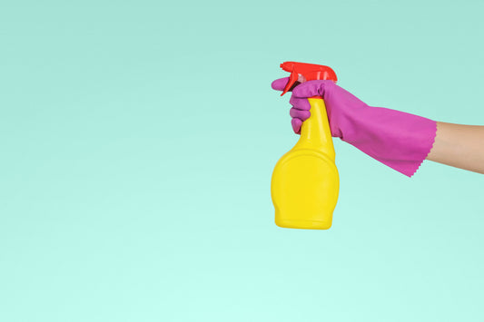 Hand with a pink cleaning glove holding a yellow and red spray bottle against a sea green background
