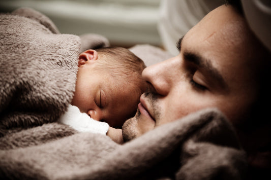 Picture of a man sleeping with a newborn baby sleeping on his chest wrapped in a brown blanket