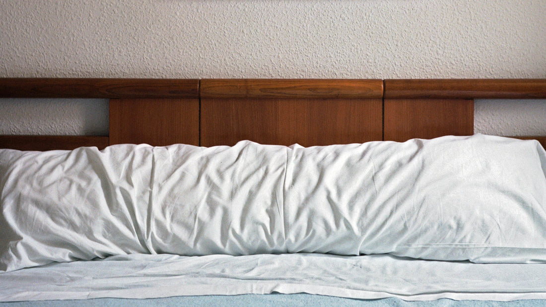 Body pillow in a white body pillowcase leaning up against a dark wood headboard