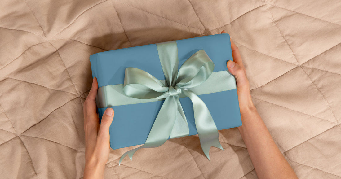 Two hands holding a wrapped gift with a bow over a Tuft & Needle Quilt