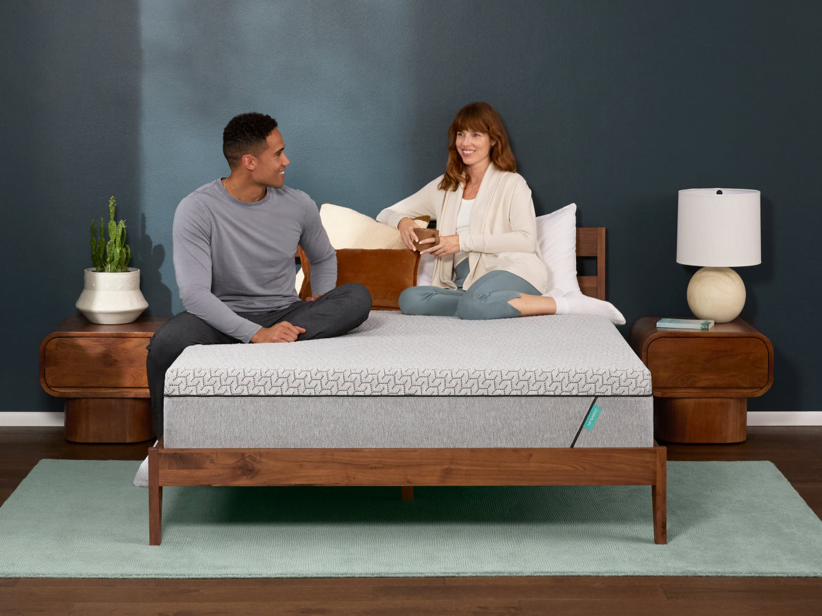 A couple relaxes on a modern bed with a light grey mattress, reading and drinking coffee in a cozy, minimalist bedroom.