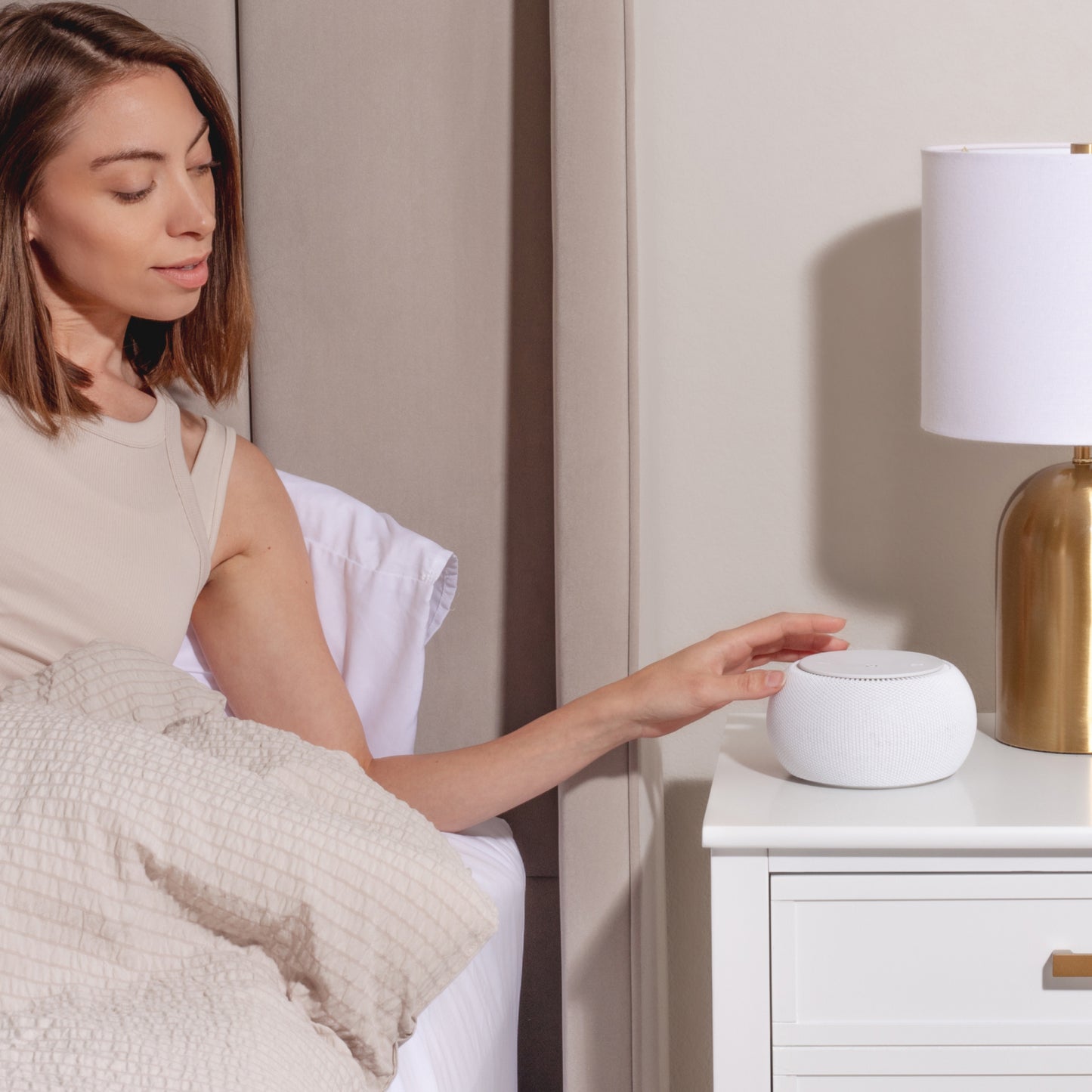 Woman sitting on a bed using a Tuft and Needle snooz pro white noise machine on a nightstand next to a gold lamp.