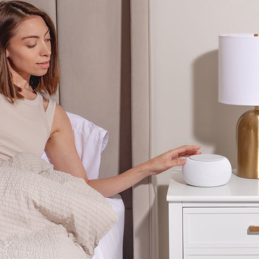 Woman sitting on a bed using a Tuft and Needle snooz pro white noise machine on a nightstand next to a gold lamp.