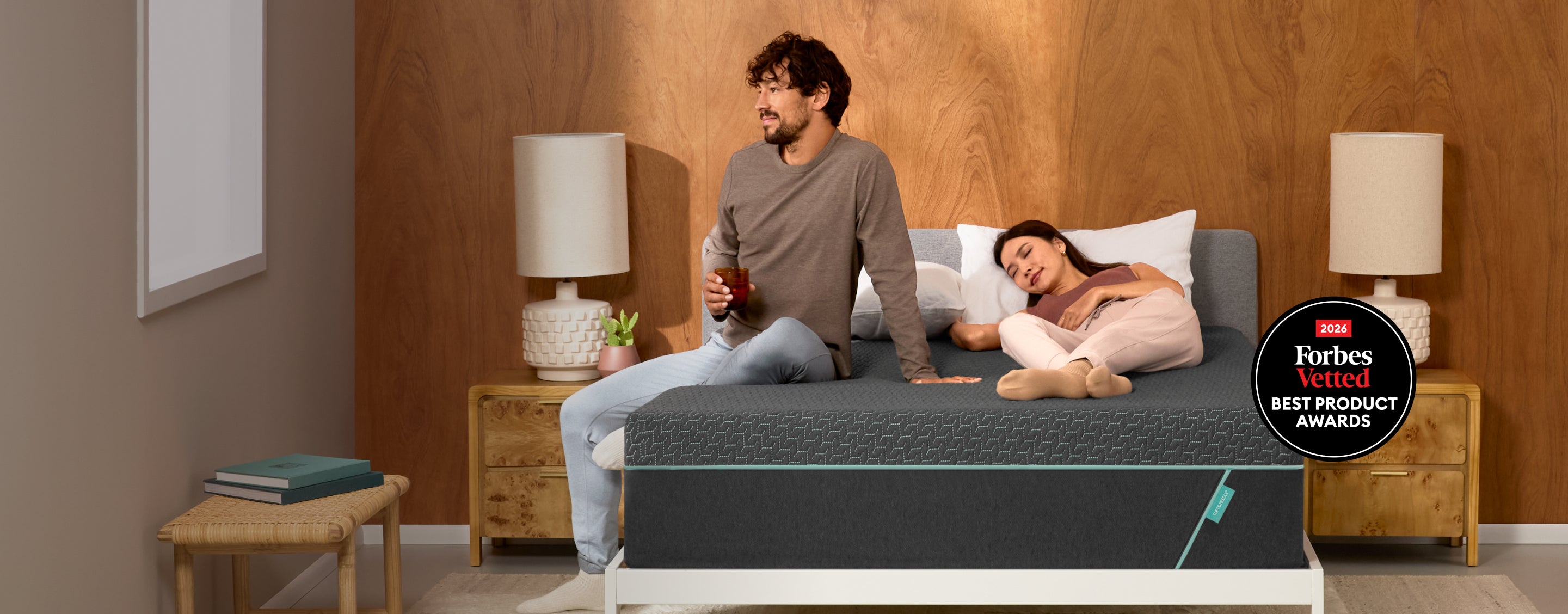 A man and a woman are shown on a Tuft and Needle Mint Hybrid mattress in a bedroom setting, with the bed mattress as the apparent product. The scene includes bedside tables, lamps, wood-paneled walls, and a circular 'Forbes Best Product Awards' badge on the right side.