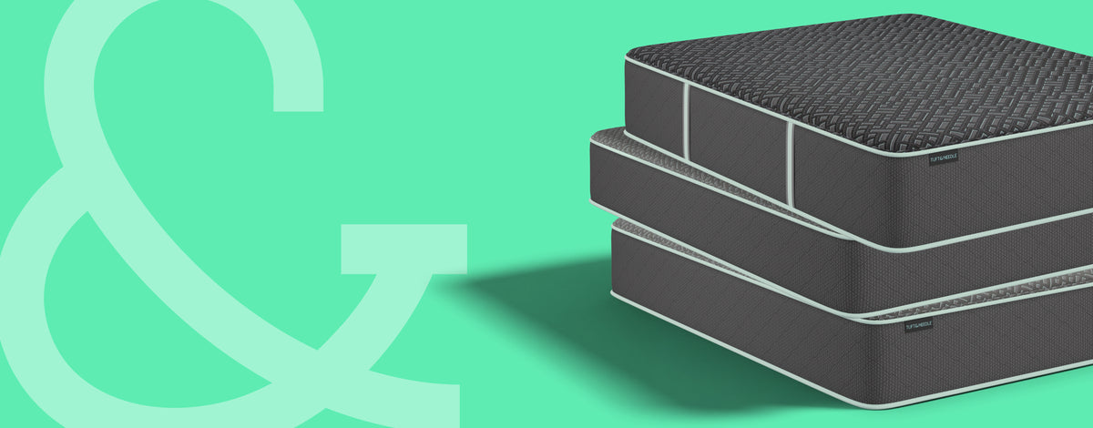 Three stacked Tuft and Needle mattresses in dark gray and black with white piping, shown on a mint green background with a large pale green ampersand graphic on the left.