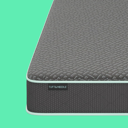 Close-up of a Tuft and Needle mattress corner showing a textured dark gray top surface and side panel with a visible TUFT & NEEDLE label, set against a solid mint green background.