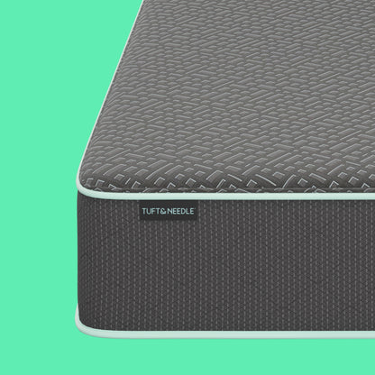 Close-up corner view of a the Tuft and Needle  13.5" Mint Hybrid II mattress showing a dark gray textured top surface and side panel with a visible TUFT & NEEDLE label, set against a solid mint green background.