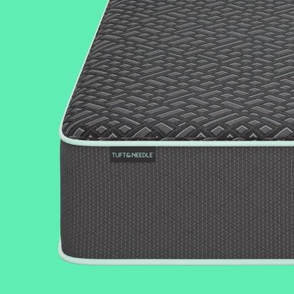 A dark gray Tuft & Needle textured mattress shown at an angle against a mint green background with abstract light green shapes. The brand name Tuft & Needle is visible on a small label on the side of the mattress.