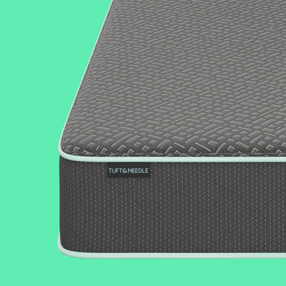 Close-up of a Tuft and Needle mattress corner showing a dark gray textured top surface, a black side label reading TUFT & NEEDLE, and a white edge trim against a bright mint green background.