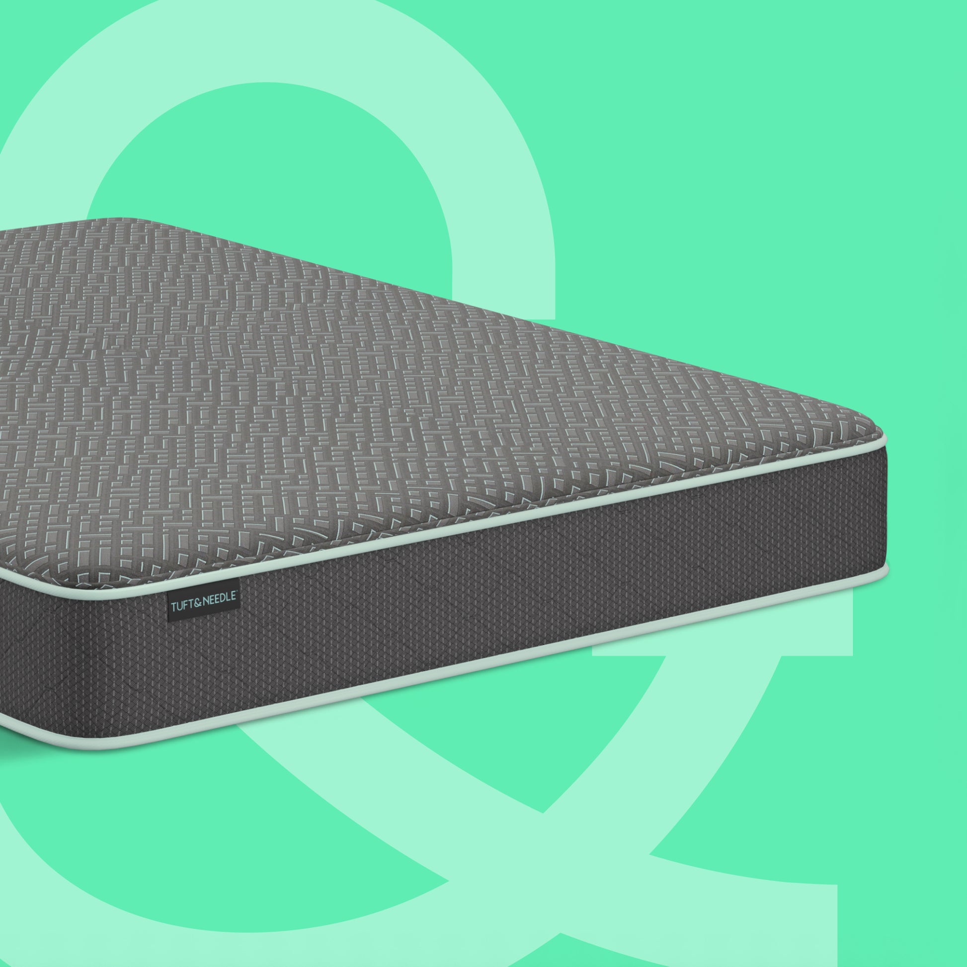 A dark gray Tuft and Needle mattress with a textured quilted top and a dark gray side panel, shown at an angle on a mint green background with abstract light green shapes.