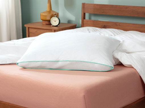 A white pillow with a mint green stitched edge resting on a tuft and needle mattress with a peach-colored sheet and white duvet, next to a wooden headboard and bedside table.