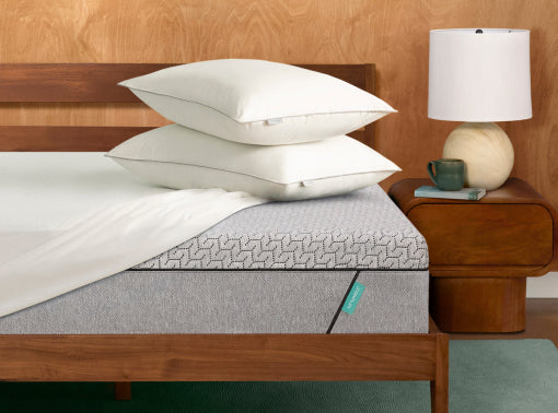 A Tuft and Needle Mint mattress with a gray patterned, white bedding, and two stacked white pillows in a bedroom setting with a wooden headboard, bedside table, and lamp.