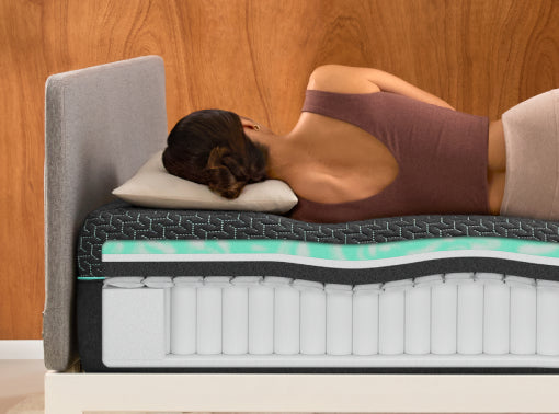 A woman lies on her side on a Tuft and Needle mint mattress with a gray upholstered headboard, shown against a wood-paneled wall. The mattress side profile reveals layered cushioning in gray, mint green, and white.