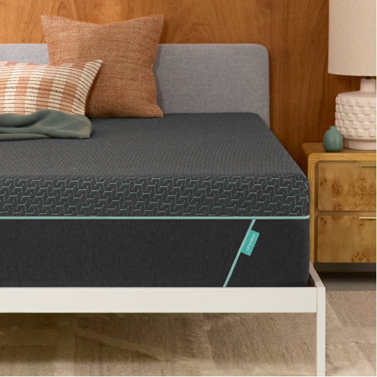Tuft & Needle Mint Hybrid mattress on a white bed frame with tan and green pillows beside a wooden nightstand.