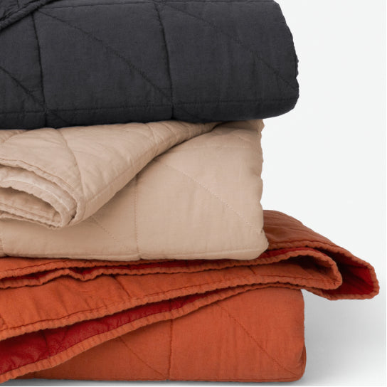 Stack of folded Tuft & needle quilted blankets in black, beige, and orange on a light gray background