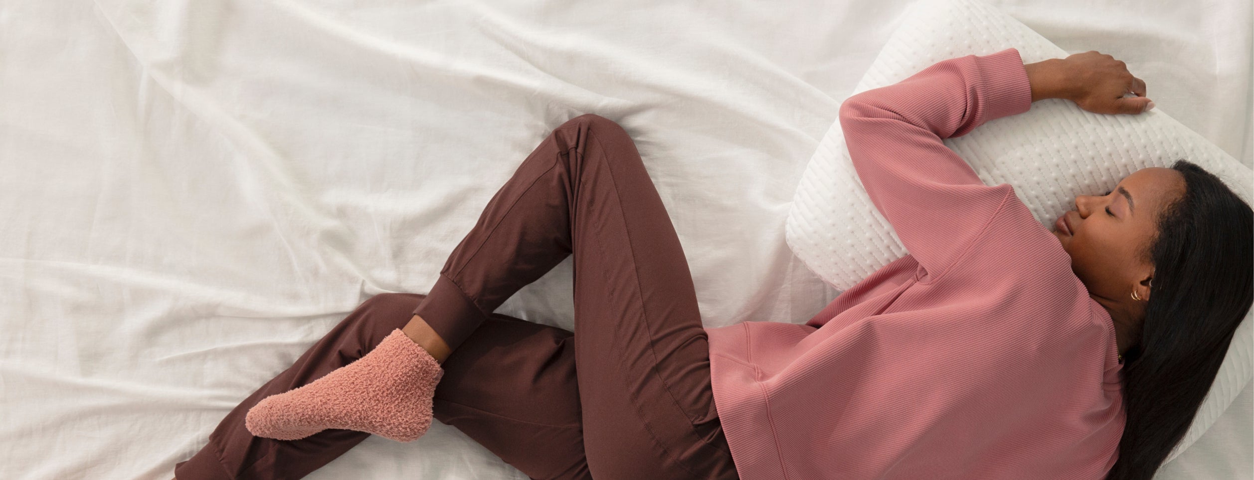 Woman lying on a Tuft and Needle mattress wearing a pink sweatshirt and brown pants with her head on a Tuft and Needle Original Foam Pillow