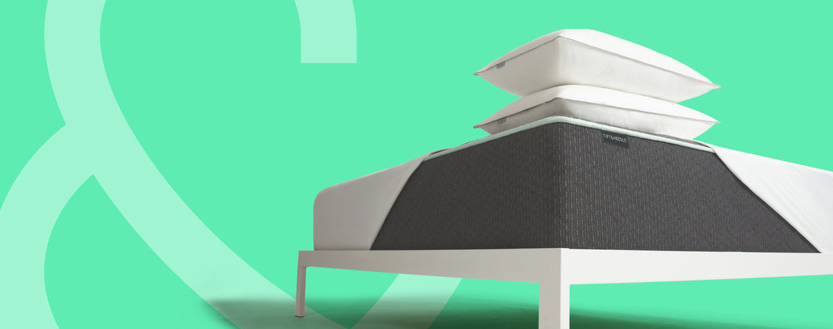A white Tuft and Needle bed frame with 2 stacked white pillows and a dark textured Tuft and Needle Original II mattress, shown against a mint green background with abstract curved line graphics.