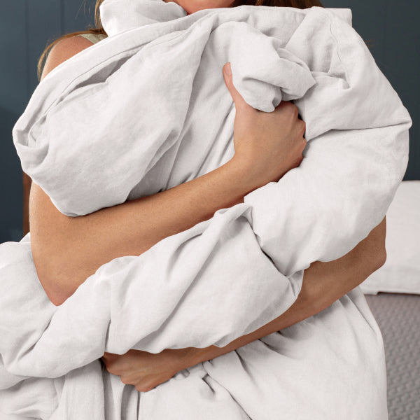 A close-up of a person hugging a light gray or off-white Tuft and Needle duvet cover and pillowcase in a bedroom setting.