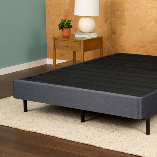 A dark gray upholstered Tuft and Needle bed frame with black legs, shown in a styled bedroom setting with a wooden nightstand, lamp, plant, and wood-paneled wall.