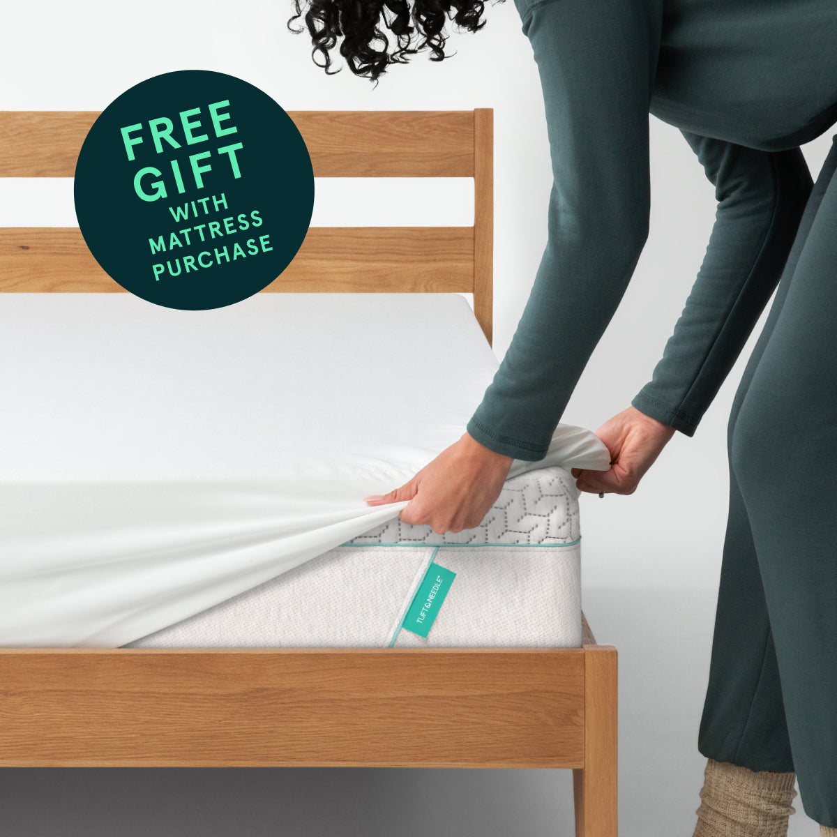 Woman standing to the right of a Tuft & Needle mattress, placing a mattress cover on  the corner of the mattress.  With a circular sticker stating Free gift with mattress purchase