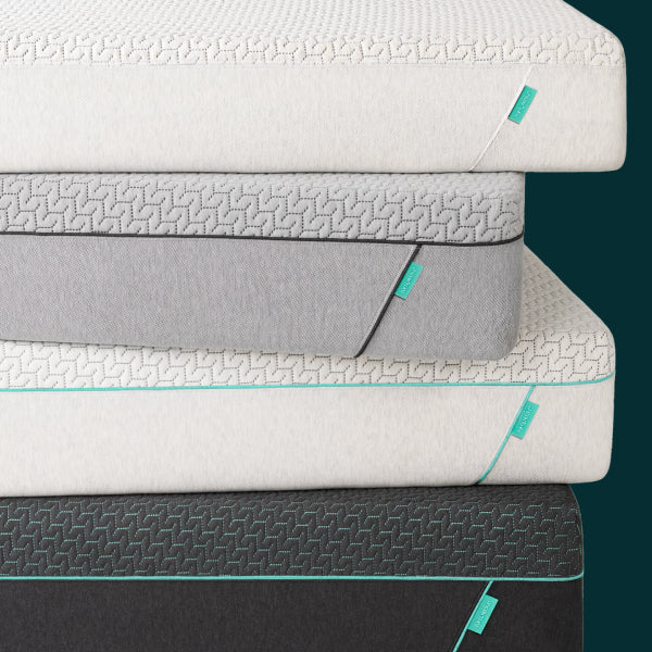Four stacked Tuft and Needle mattresses, Original, Original Hybrid, Mint and Mint Hybrid in different colors and textures, including white, gray, and dark charcoal, with small teal brand tags visible on the side.