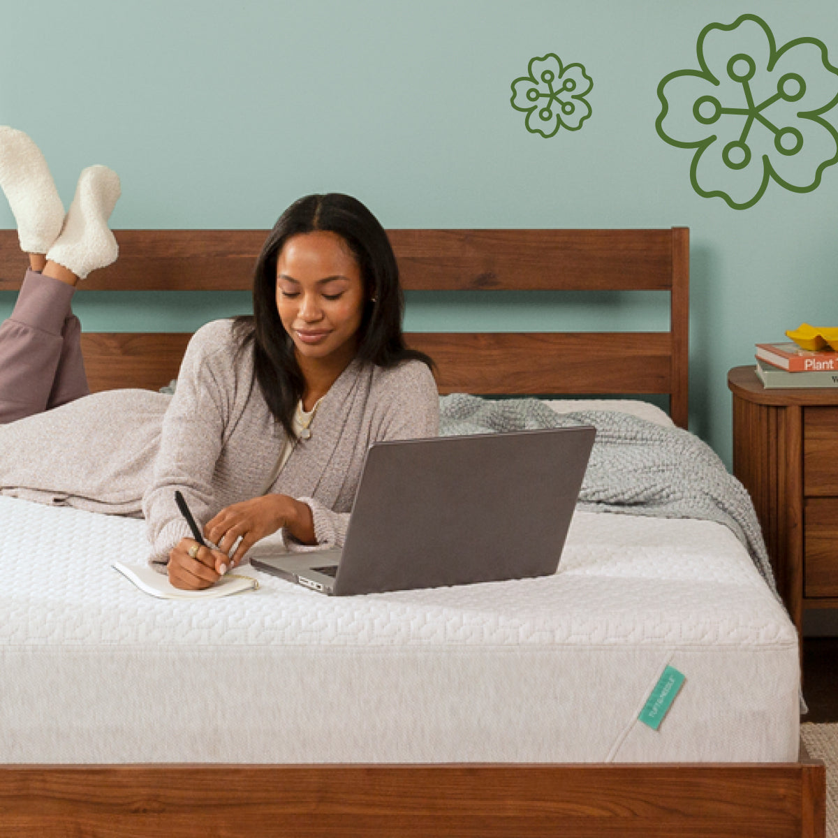 Woman using a laptop on a Tuft and Needle Original mattress with a visible mattress brand tag, against a light blue wall with green floral designs.